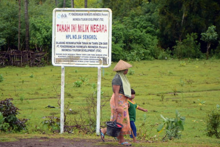Seorang perempuan berjalan di dekat papan yang menunjukkan kawasan ini milik negara dalam kelola PT ITDC. Banyak petani di Desa Kuta dan Desa Sengkol becocok tanam di atas lahan ITDC. Foto: Fathull Rakhman / Mongabay Indonesia