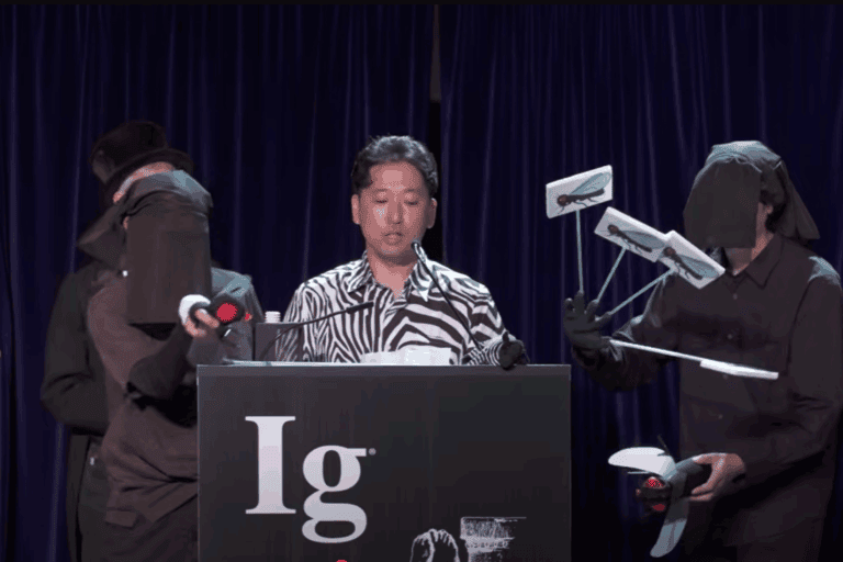 Tomoki Kojima, peneliti Jepang, tampil dengan kemeja bermotif zebra saat menerima Ig Nobel 2025 kategori biologi. Rekannya memperagakan “serangan lalat” di panggung sebagai bagian dari presentasi humoris mereka | Sumber: Improbable Research / YouTube (tangkapan layar)
